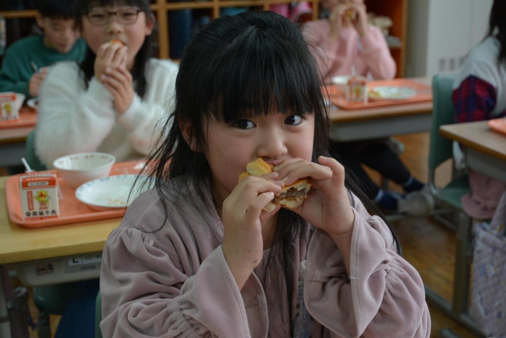 写真：赤身と脂のバランスがいい秋田由利牛と野菜を、パンにはさんだサンドイッチを食べる児童2