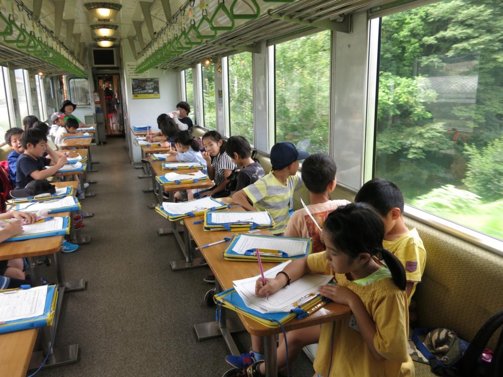 写真：列車内で子吉川や鳥海山の景色を楽しんだり，おばこアテンダントから教えていただいたことをメモする児童