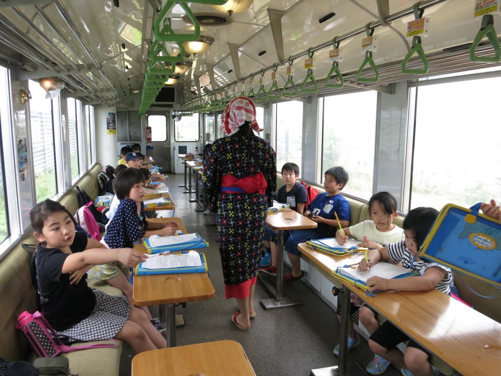 写真：列車内で子吉川や鳥海山の景色を楽しんだり，おばこアテンダントから教えていただいたことをメモする児童2